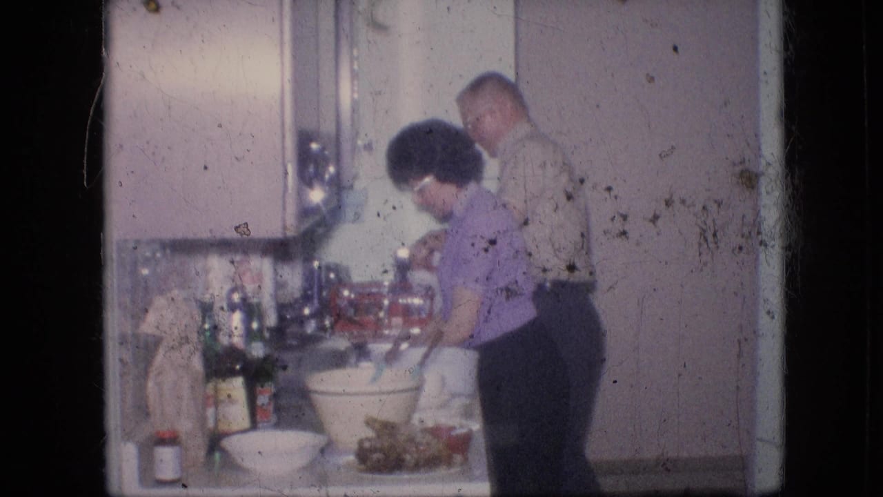 In a 1964 Philadelphia home-movie frame, an older woman and older man appear to prepare supper at a kitchen counter with bowls, bottles, and white cabinets nearby.