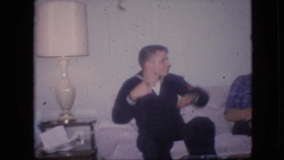 A young man in a dark uniform-like top sits on a sofa in a Philadelphia living room, with a table lamp and another seated person partly visible.