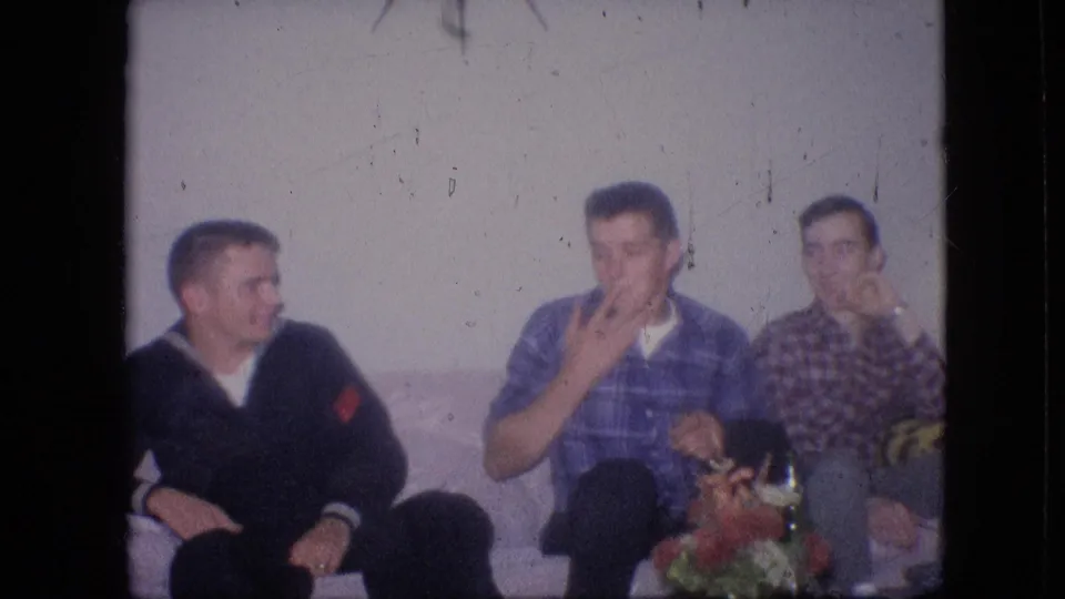 Three young men sit closely on a sofa in a 1964 Philadelphia home interior, including one in a dark uniform-like top and two in plaid shirts.