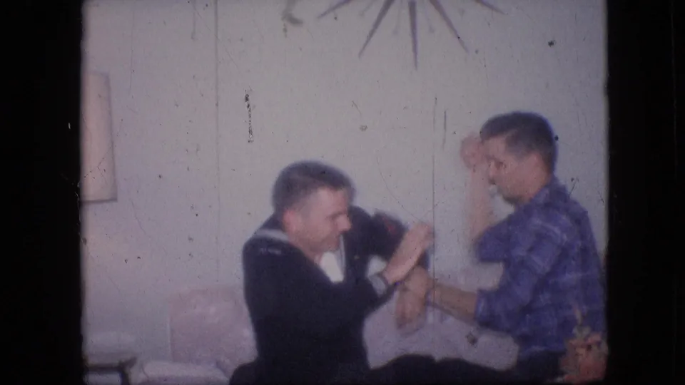 Two young men playfully interact on a sofa in a 1964 Philadelphia living room, with a starburst wall decoration and soft home-movie focus visible.