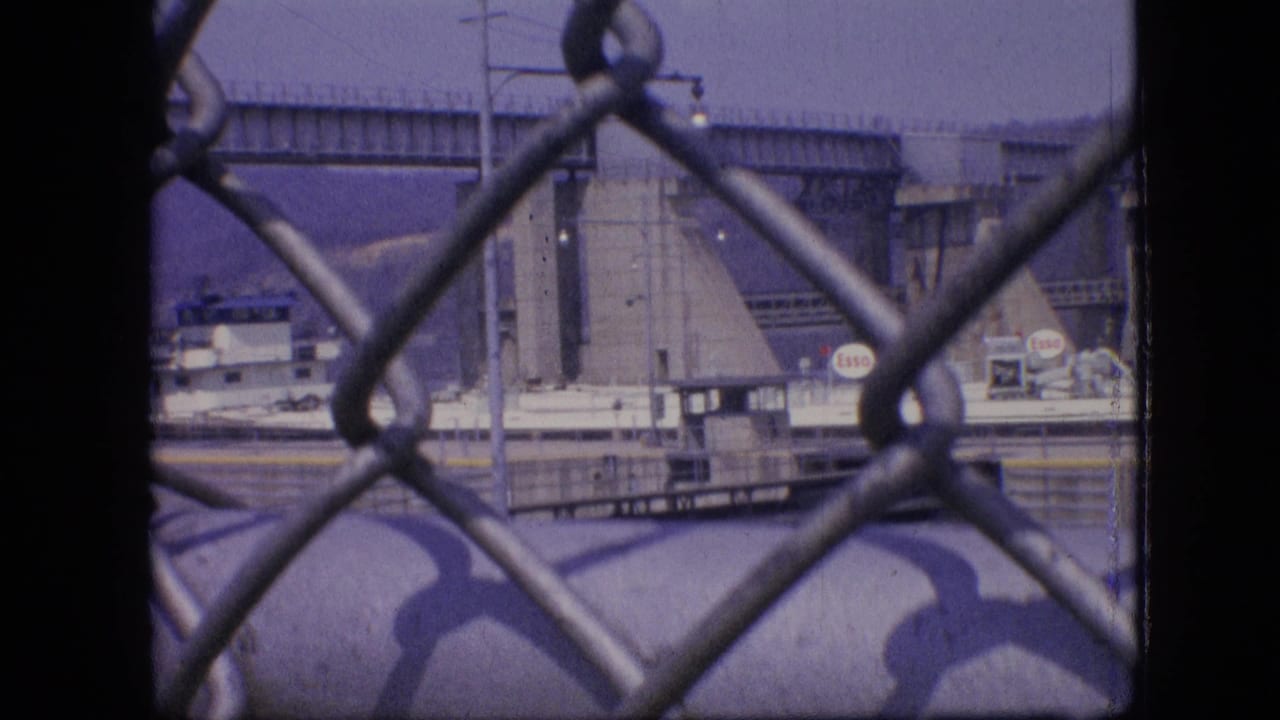 1964 Philadelphia waterfront view through a chain-link fence, with a docked industrial vessel, bridge structure, and Esso signage in the distance.