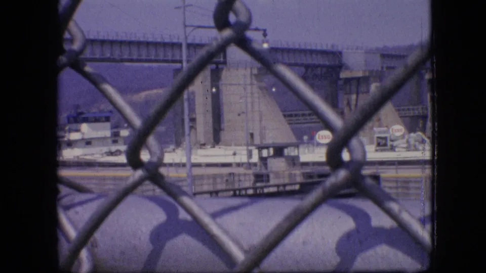 1964 Philadelphia waterfront view through a chain-link fence, with a docked industrial vessel, bridge structure, and Esso signage in the distance.