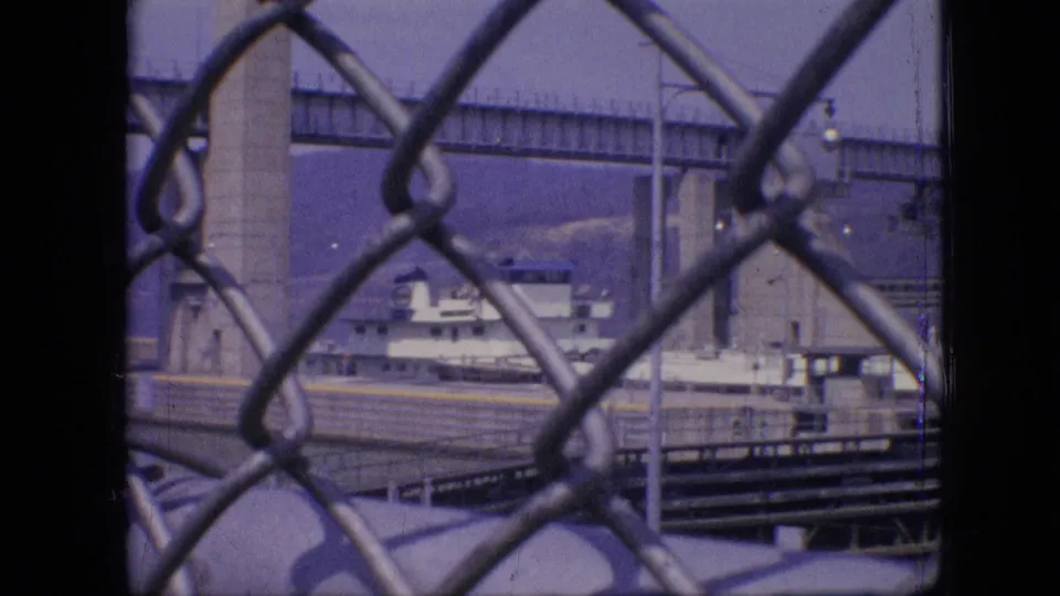Philadelphia industrial waterfront scene with chain-link fencing in the foreground, a large vessel at dock, bridge piers, and visible Esso branding.