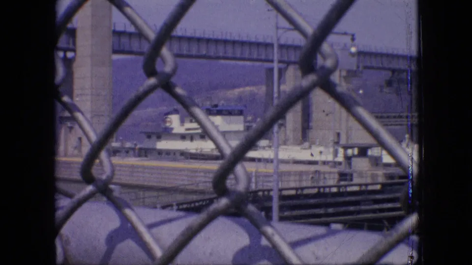 Home-movie frame of a Philadelphia waterfront shipyard area, showing fence links in front of a vessel, dock railings, and a bridge support.