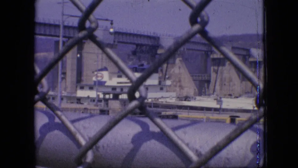 1964 Philadelphia shipyard view through chain-link fencing, with the vessel, bridge span, concrete pier, and waterfront equipment visible under a hazy sky.
