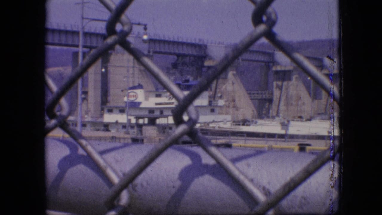 1964 Philadelphia home-movie frame showing a white and blue boat in a concrete lock or canal system, viewed through a chain-link fence.