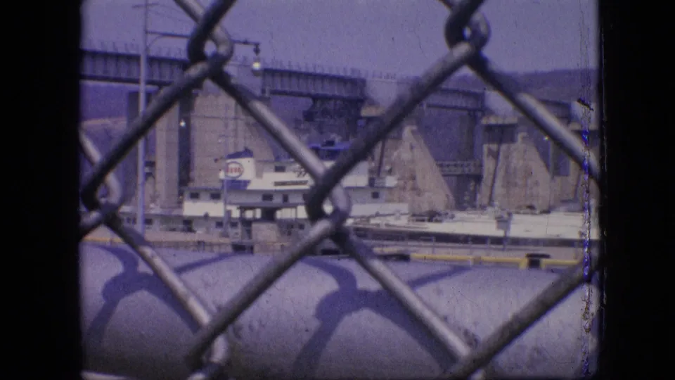 1964 Philadelphia home-movie frame showing a white and blue boat in a concrete lock or canal system, viewed through a chain-link fence.