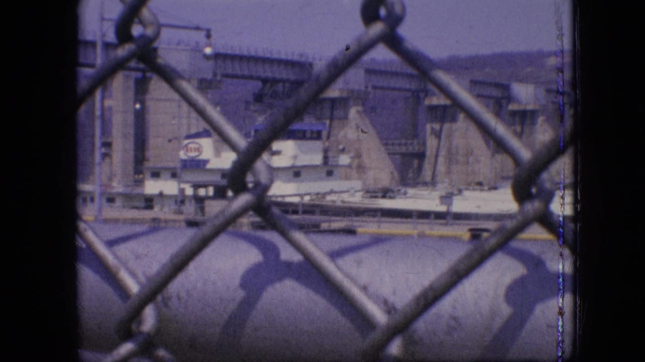 1964 Philadelphia waterway footage frame with a vessel passing concrete lock infrastructure while foreground fence links dominate the view.