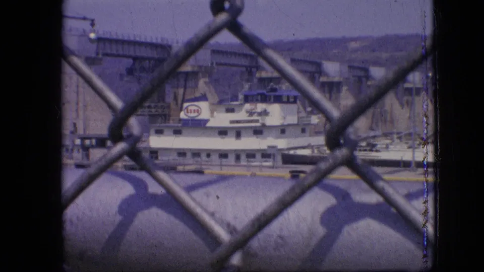 Final frame from the 1964 Philadelphia clip showing the white and blue boat aligned with concrete lock walls, partially obscured by chain-link fencing.