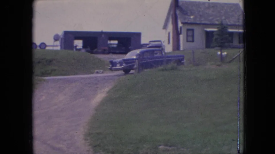 A blue sedan moves across the frame near a farmhouse, barn or garage, grassy slope, and open rural roadside in the supplied 1964 Pennsylvania clip.