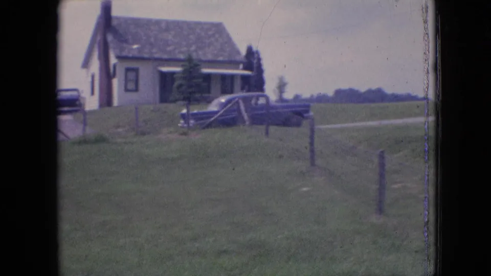 The blue sedan is partly side-on near a farmhouse and rural roadside, with fence posts and open fields visible in the soft 8mm frame.