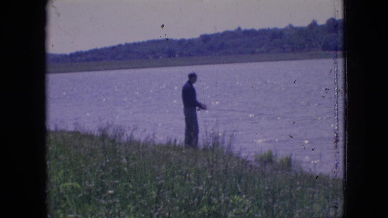A distant man in dark clothing stands on a grassy lakeside bank with a fishing rod in 1964 Philadelphia-area home-movie footage.