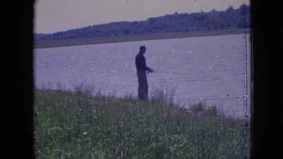 A lone fisherman stands near the edge of a lake, with open water and a wooded far shore visible in archival 8mm footage from Philadelphia, Pennsylvania.