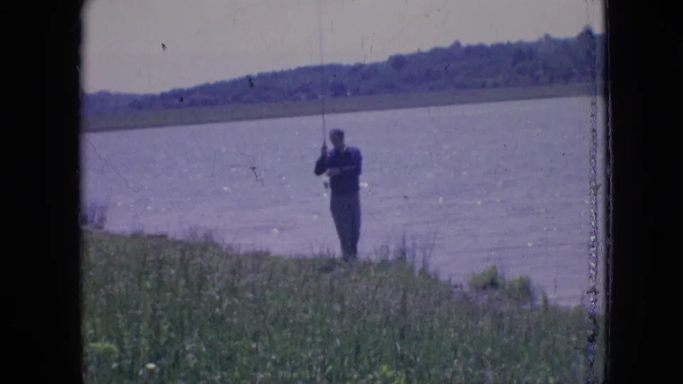 The fisherman lifts his rod near the lakeshore, framed by grass in the foreground and a wide body of water behind him in 1960s 8mm home-movie footage.