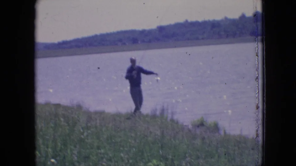 A man by the water extends one arm while handling a fishing rod on a grassy bank, with the lake and distant wooded shore behind him.