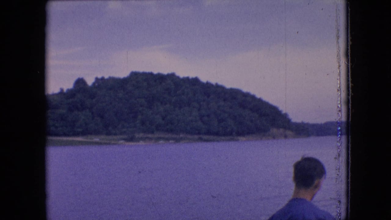 1964 8mm home-movie frame from the Philadelphia metadata context showing a man from behind at the water's edge with wooded hills across the lake.