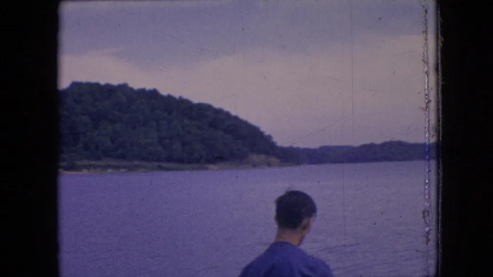 Metadata-supported 1964 Philadelphia-area home-movie frame with a man turned away from the camera, open water, and a wooded shoreline in the distance.