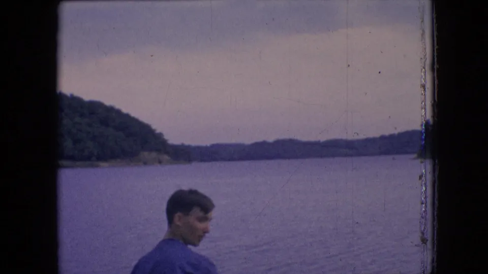 1964 8mm frame from the Philadelphia metadata context showing a man in partial profile near the water, with low wooded hills and an overcast sky behind him.