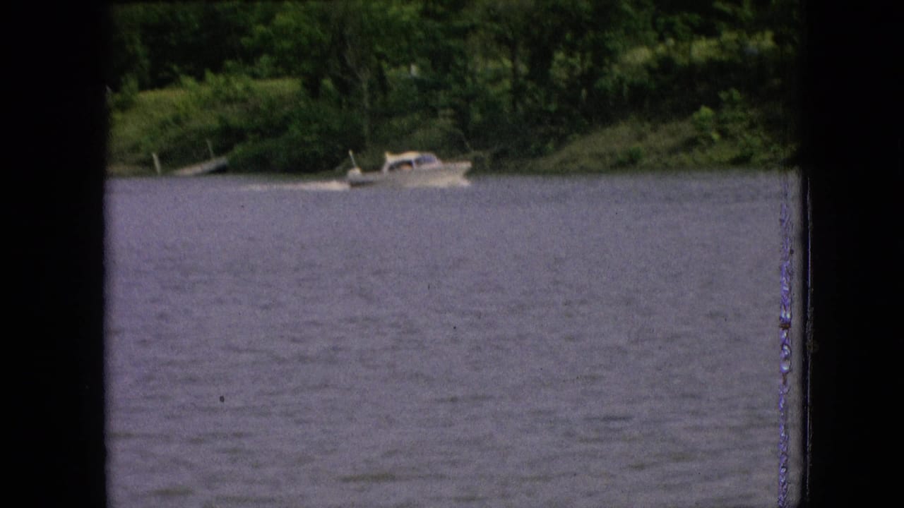 1964 archival 8mm frame showing a small boat near a tree-lined shore across a broad, calm body of water, with visible film gate edges and grain.