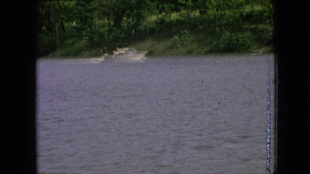 Archival 8mm view of a small boat along a wooded shoreline, with the water filling most of the frame and dark contact-sheet borders visible.