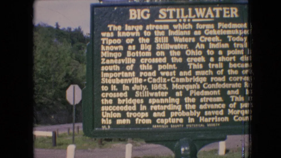 1964 archival 8mm frame showing a green Big Stillwater historical marker near a road, with trees, roadside posts, and the back of a stop sign visible behind it.