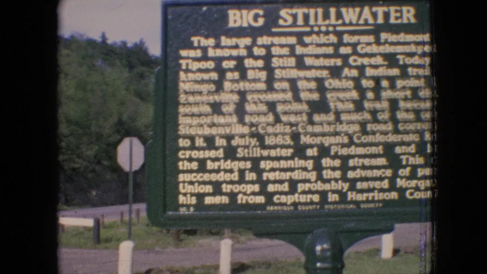 Archival 8mm close view of the Big Stillwater roadside historical marker, with dense text, a wooded background, white posts, and a road edge visible.