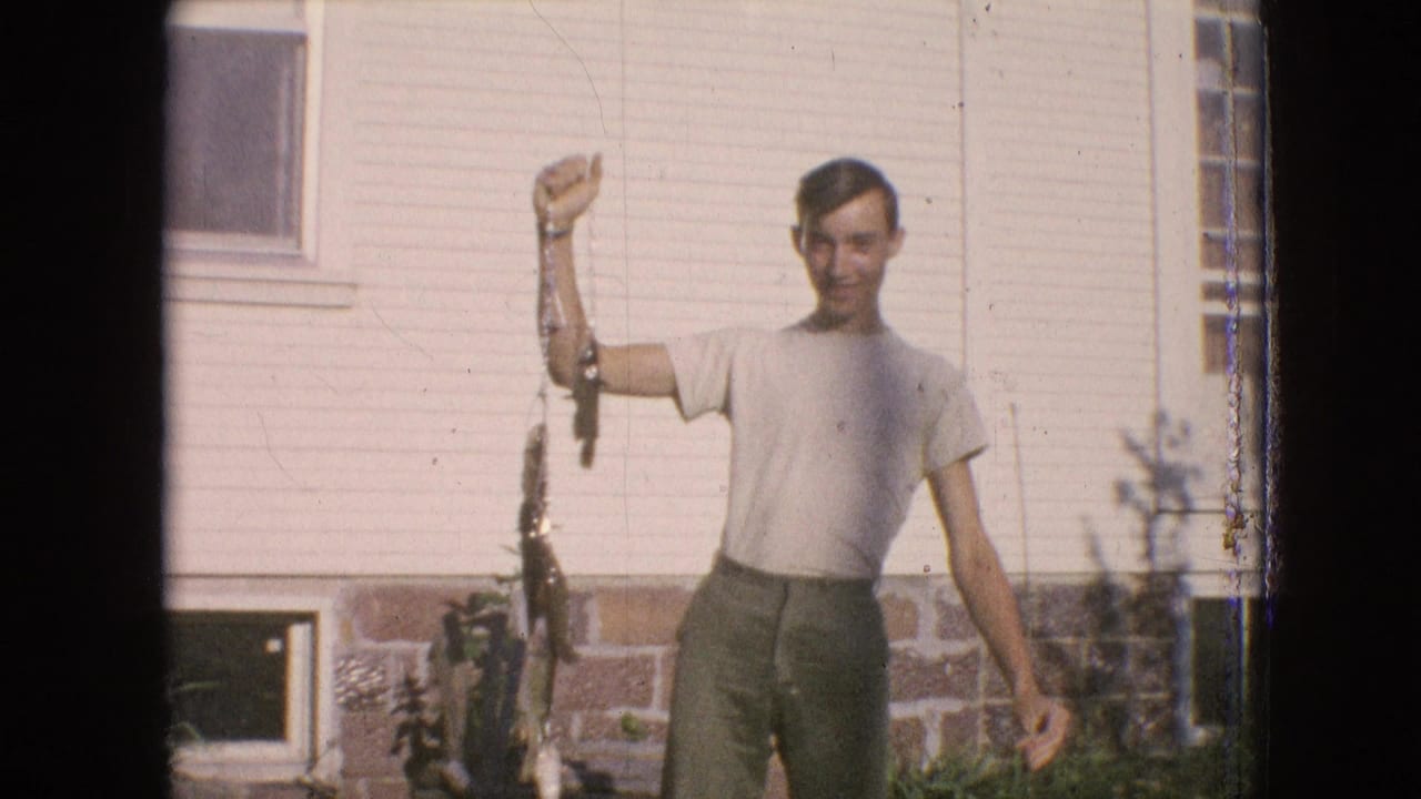 In 1964 Philadelphia home-movie footage, a young man in a light T-shirt and dark trousers smiles while raising a line of fish in a grassy yard beside a house with siding and a brick foundation.