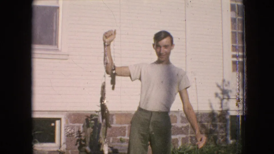 In 1964 Philadelphia home-movie footage, a young man in a light T-shirt and dark trousers smiles while raising a line of fish in a grassy yard beside a house with siding and a brick foundation.