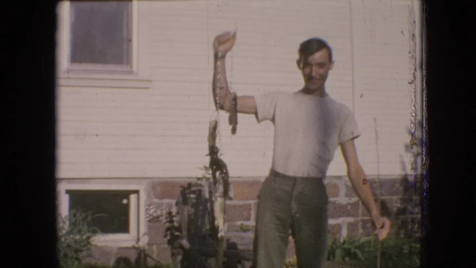 The young man stands in bright sunlight in a Philadelphia residential yard, holding a fish line above his shoulder while facing the home-movie camera.