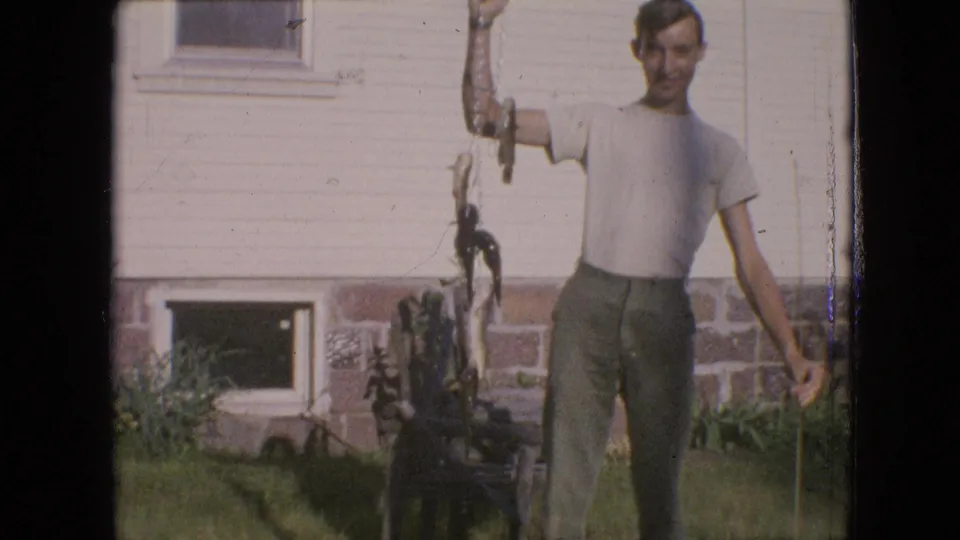 A closer handheld frame shows the young man and dangling fish against the house exterior, with the grassy yard and small basement window visible below.