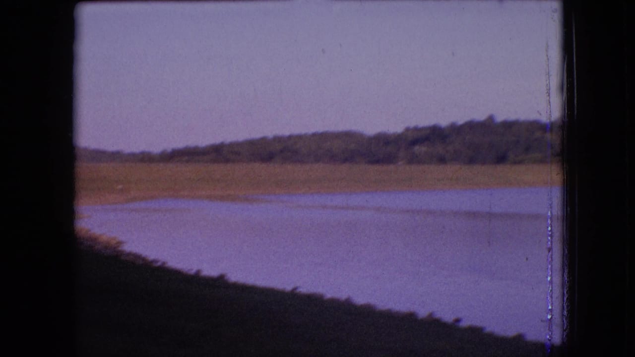 1964 archival 8mm frame showing a calm lake near Philadelphia, Pennsylvania, with a low shoreline, wooded ridge, pale sky, and dark foreground bank.