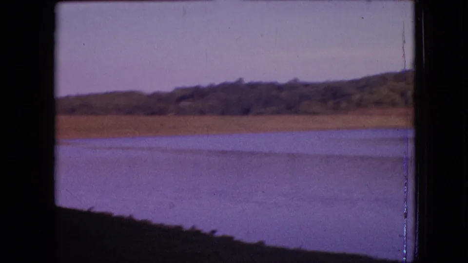 1964 archival 8mm frame of a still lakeside view in the Philadelphia, Pennsylvania metadata location, with sandy shoreline, distant trees, and purple-shifted water tones.