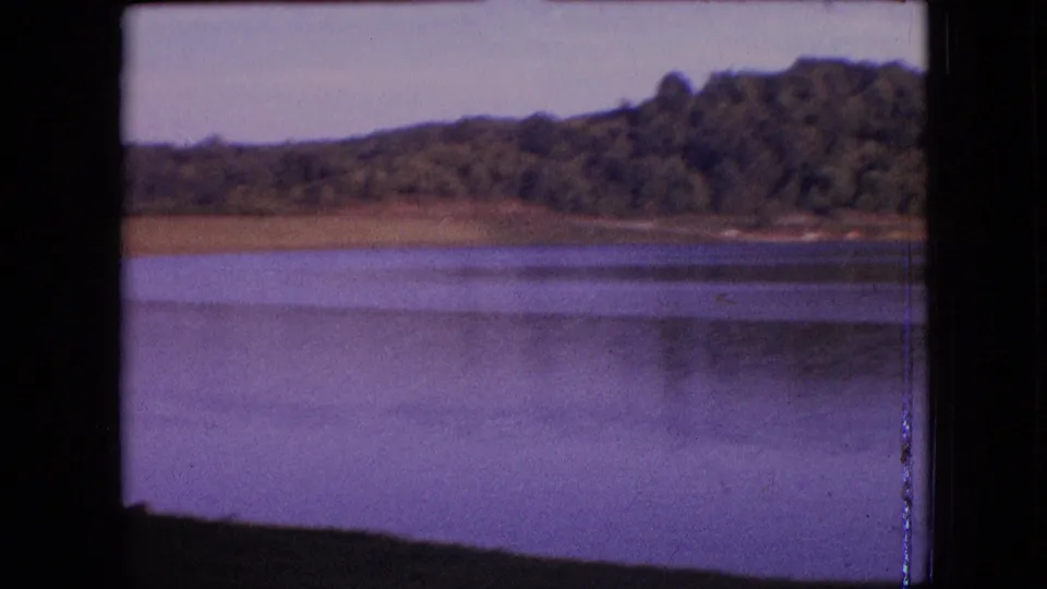 1964 archival 8mm frame showing lake water with faint reflections, a narrow shoreline, and dense trees on a low hillside.