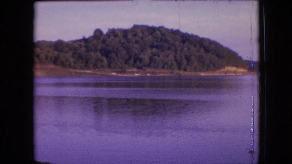 1964 archival 8mm frame of a forested hill across calm water, with a narrow exposed shore and visible film-edge wear.