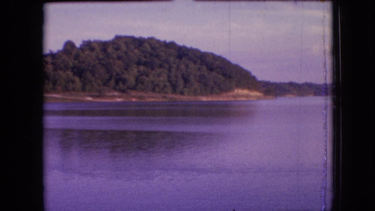 1964 metadata-supported 8mm frame of a calm lake near Philadelphia, Pennsylvania, with a tree-covered hill, distant shore, soft sky, and visible film-edge wear.