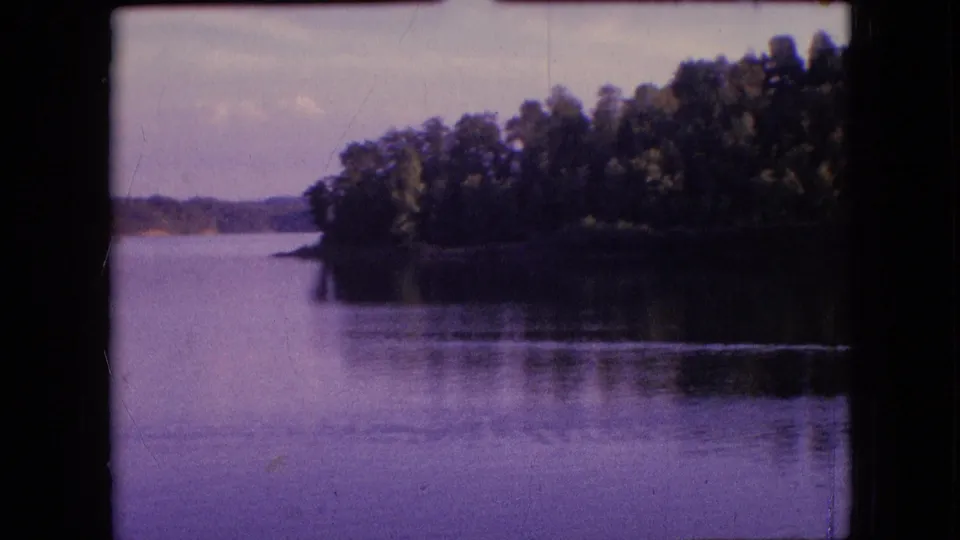 1964 metadata-supported 8mm frame of calm water near Philadelphia, Pennsylvania, with a shaded wooded shoreline to the right and low hills in the distance.