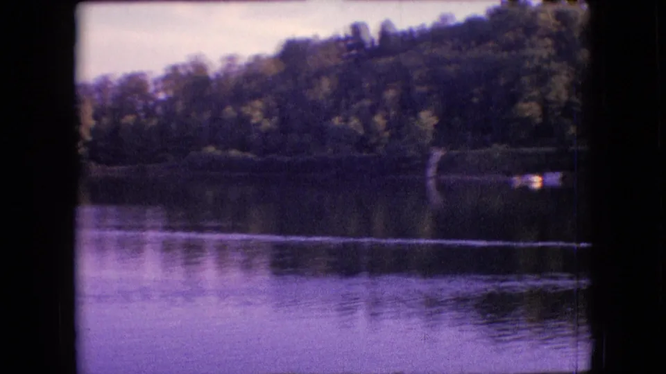 1964 metadata-supported 8mm frame near Philadelphia, Pennsylvania, showing a tree-lined lakeshore, reflective water, hillside vegetation, and a small bright object at the far right edge.