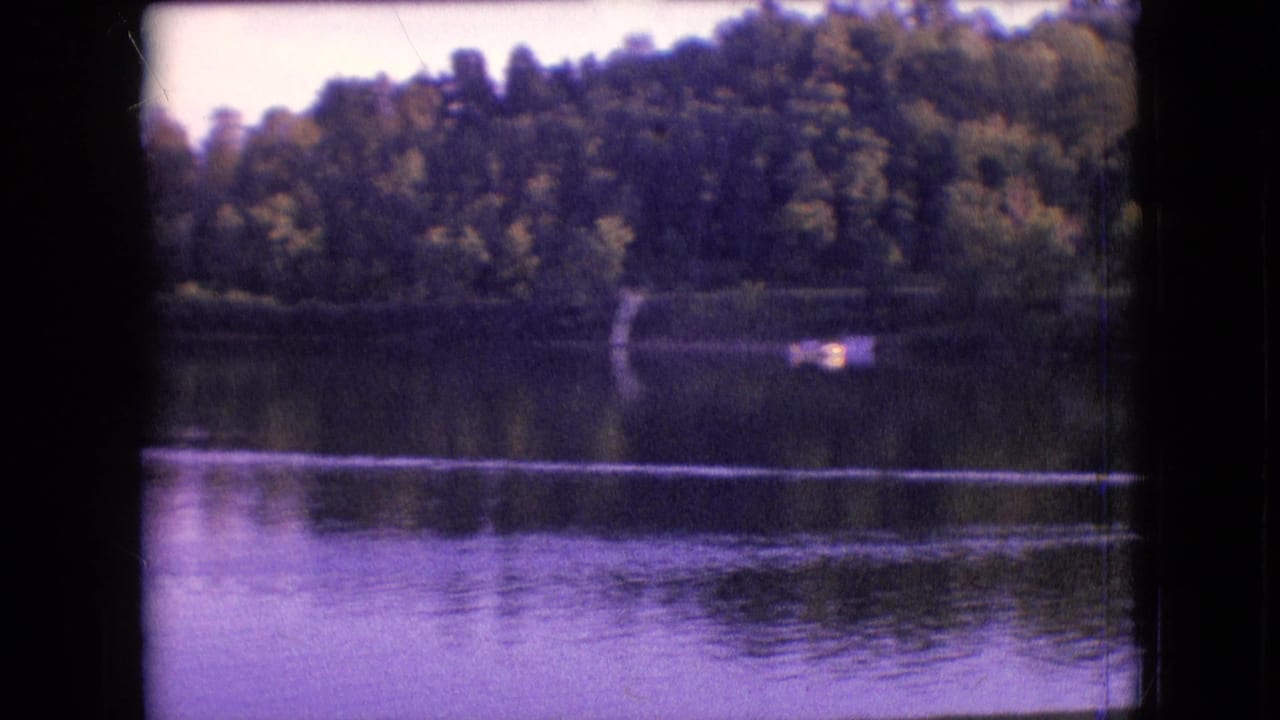 1964 Philadelphia-area 8mm frame showing a calm lake, tree-lined shore, water reflections, and a small distant boat moving across the water.