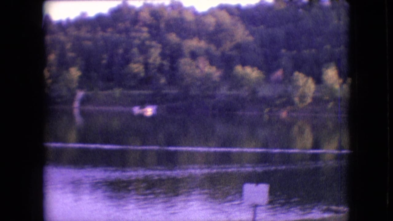1964 Philadelphia-area 8mm frame of a distant boat on a calm lake, with wooded shoreline reflections and a white signpost near the lower right edge.
