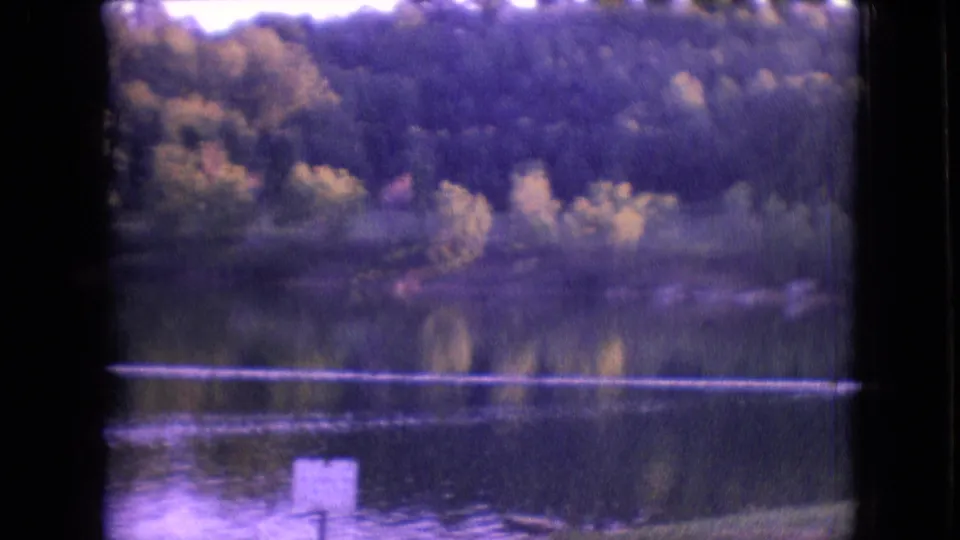 1964 Philadelphia-area 8mm frame with a calm lake, reflected trees, wooded bank, and a white lakeside sign in the lower foreground.