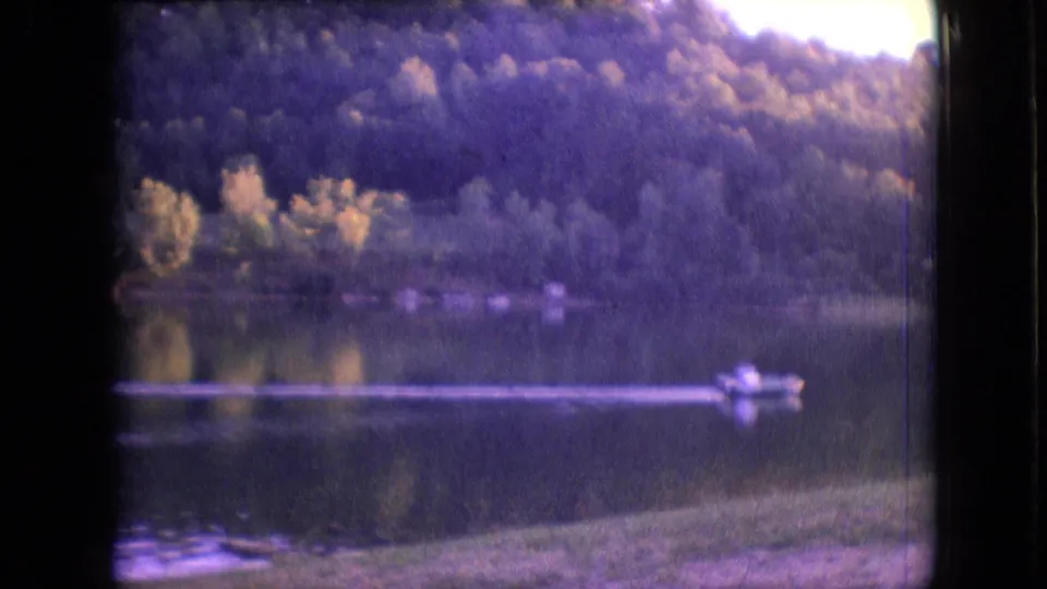 1964 Philadelphia-area 8mm frame showing a small boat with indistinct occupants near the right side of a calm lake, backed by trees and a grassy shoreline.