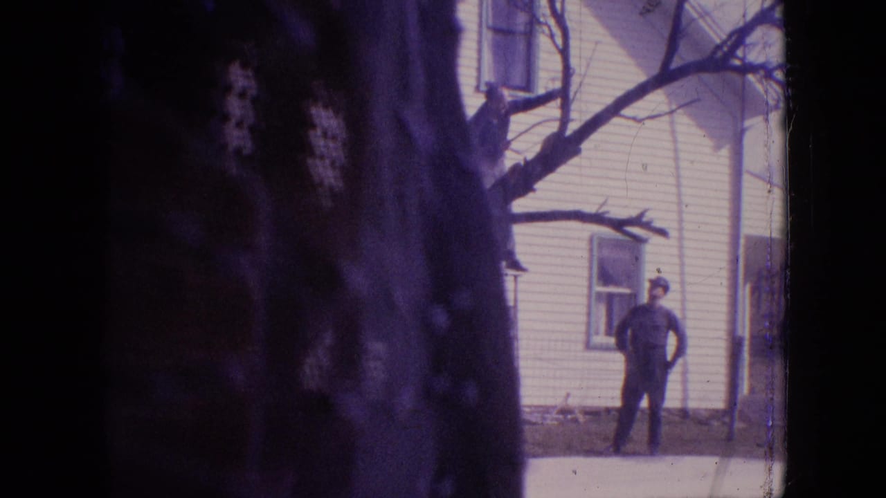 Man standing near a white house while a large tree trunk frames the foreground.