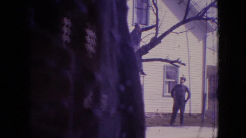 1964 Philadelphia home-movie frame showing a man standing near a white two-story house, with a large dark tree trunk and branch dominating the foreground.