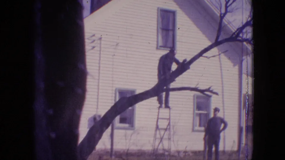 1964 Philadelphia home-movie frame showing a man on a ladder cutting or handling a branch in front of a white two-story house while another man stands below.