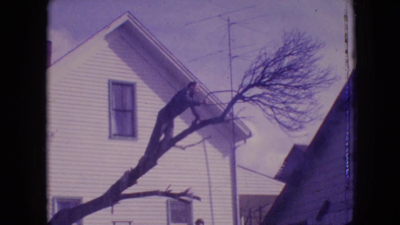 1964 Philadelphia home-movie frame showing a man in dark clothing positioned on a large bare tree limb beside a white house.