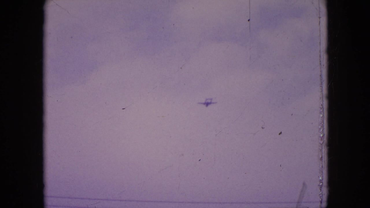 A distant small propeller plane appears against a cloudy sky in 1964 Philadelphia 8mm home-movie footage, framed by dark film edges and visible surface wear.
