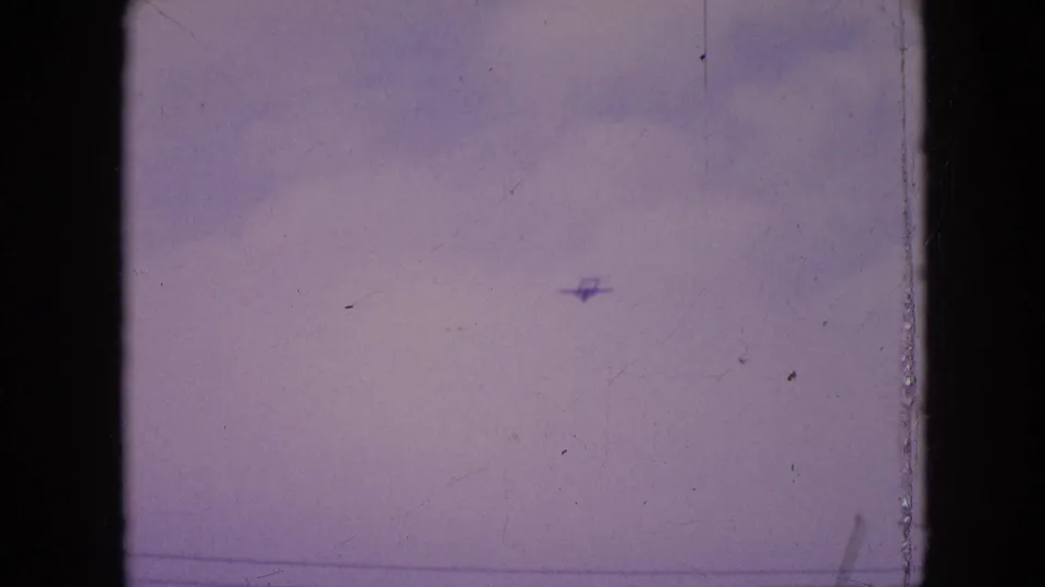 A distant small propeller plane appears against a cloudy sky in 1964 Philadelphia 8mm home-movie footage, framed by dark film edges and visible surface wear.
