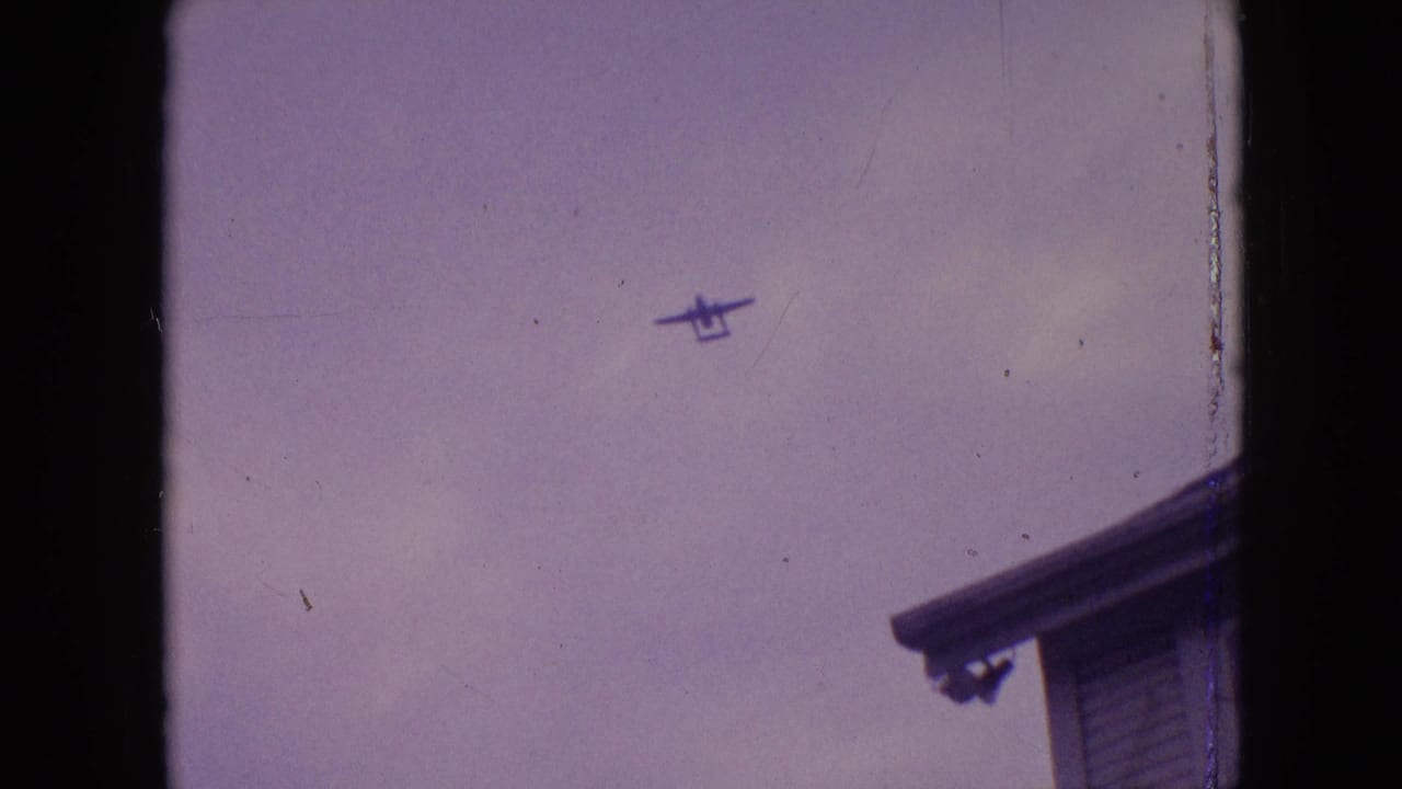 Small plane high in a cloudy sky with a roof edge at lower right.