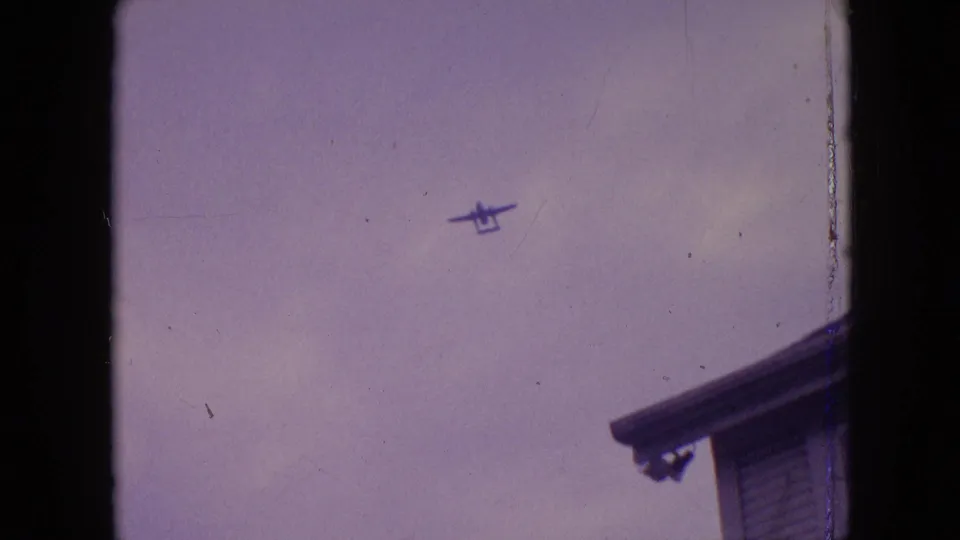 A small propeller plane is seen in the cloudy sky above Philadelphia, with a roof edge entering the lower right of the 8mm frame.
