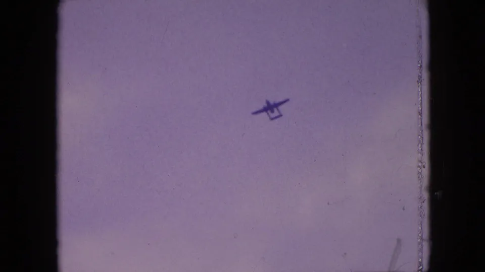 The small propeller plane appears slightly lower and larger in the 8mm frame, set against a pale cloudy sky with visible film wear.